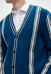 Μπλε μάλλινο ζακετάκι με λευκές και μαύρες ρίγες. Διαθέτει V-λαιμόκοψη, πέντε κουμπιά και υφή με ανάγλυφο φινίρισμα. Λευκό πουκάμισο από κάτω.