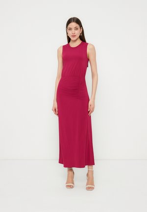 Fiatal nő áll, ujjatlan, földig érő fukszia ruhát visel, összehúzott derékdísszel és bézs színű nyitott orrú magas sarkú cipővel, fehér háttér előtt.