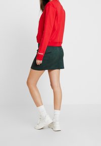 Röd sweatshirt med rund halsringning, matchad med en mörkgrön mockakjol. Modellen har på sig vita ankelsockar och vita sneakers.