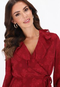 Czerwona kwiecista sukienka kopertowa z falbanką przy dekolcie, fakturowaną tkaniną i wiązaniem w talii. Złote kolczyki koła dopełniają całość stylizacji.