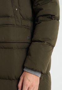 Olivengrøn pufferjakke med quiltet design, trykknaplukning og en grå manchet, der stikker ud fra ærmet.