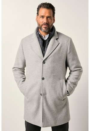 Partainen mies pukeutuneena vaaleanharmaaseen napitettuun villatakkiin mustan toppatun takin ja valkoisen paidan päällä, kädet takin taskuissa, neutraali tausta.