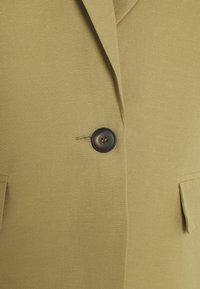 Bouton marron foncé unique attaché sur un blazer vert olive clair avec un revers cranté et deux poches à rabat.