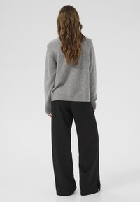 Siv svilen pulover z teksturo, sproščenega kroja in rebrastimi manšetami, v kombinaciji z ohlapnimi črnimi hlačami z bočnimi razporki. Pogled zadaj.