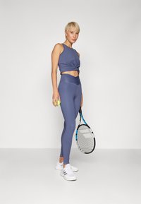 Outfit sportivo con un top senza maniche annodato di colore blu scuro e leggings a vita alta coordinati. Gli accessori includono una racchetta da tennis e una pallina.