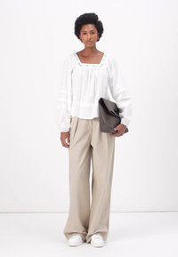 Blusa branca de mangas longas com bordado verde na gola, combinada com calças largas beiges e ténis brancos. Segura uma bolsa escura.