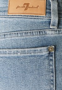 Jasnoniebieskie dżinsy mają skórzaną metkę z wytłoczonym logo oraz metalowy guzik na przedniej kieszeni.