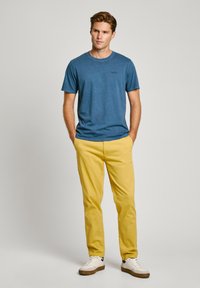 Μπλε βαμβακερή μπλούζα με κοντά μανίκια και λογότυπο στο στήθος, συνδυασμένη με κίτρινες chinos και λευκά αθλητικά παπούτσια με καφέ λεπτομέρειες.