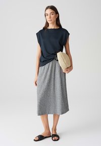 Marineblaue Kurzarmbluse, grauer strukturierter Midirock, schwarze Riemchensandalen und eine gewebte beigefarbene Handtasche. Einfaches, modernes Outfit mit neutralen Farben.