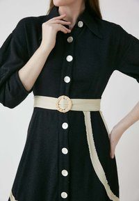 Schwarzes, strukturiertes Kleid mit Knopfleiste, übergroßem Kragen und einem beigen Gürtel mit gewebtem Ringakzent.
