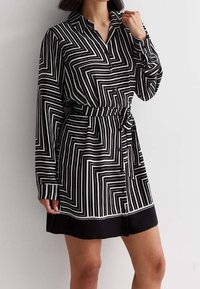 Donna che indossa un vestito camicia a maniche lunghe con stampa geometrica a righe bianche e nere, dotato di cintura e orlo nero, in piedi contro uno sfondo chiaro e uniforme.