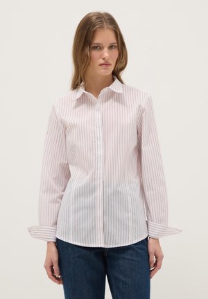 Frau trägt ein weißes Langarmhemd mit dünnen roten vertikalen Streifen und eine dunkelblaue Jeans, steht vor einem schlichten hellen Hintergrund.