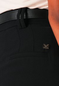 Juodos kelnės su lygiu paviršiumi, išsiskiriančios elegantišku juosmeniu, siuvinėtomis detalėmis ir metaliniu „M“ logotipu ant kišenės.