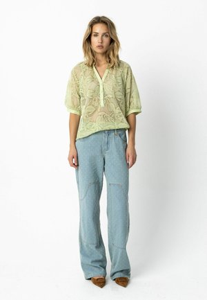 Hellgrünes, transparentes Oberteil mit Blattmuster und Knopfleiste, kombiniert mit hellblauen Jeans mit Steppnähten und Taschenakzenten.