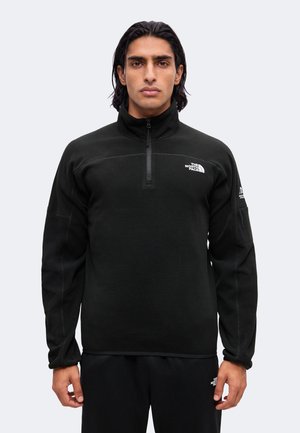Homme aux cheveux foncés portant une veste polaire noire zippée et un pantalon assorti avec des logos blancs sur la poitrine et la manche, debout devant un fond uni.