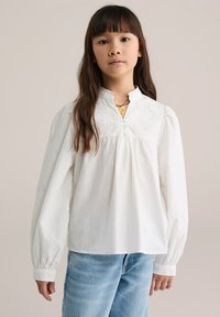 Bílá bavlněná halenka s volánovým límcem, vyšívaným předním dílem a zapínáním na knoflíky. Dlouhé rukávy s nařasenými manžetami. Nositelná s světle modrými džínami.