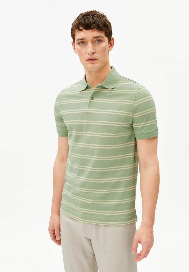 Jeune homme aux cheveux courts et bouclés portant un polo rayé vert à manches courtes et un pantalon beige, debout devant un fond blanc.