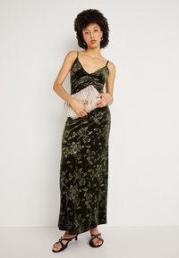 Zelené zamatové maxi šaty s kvetinovým vzorom, výstrih do V a tenkými ramienkami. Model drží svetlosivú plisovanú kabelku a má na sebe čierne sandále na opätku.