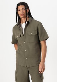 Chemise verte olive à manches courtes, en tissu léger, dotée de deux poches avant, d'un design à bouton et d'une coupe décontractée.