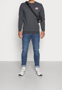 Sudadera gris oscuro con cuello redondo y puños acanalados, combinada con vaqueros azules. Pequeño logo en el pecho, bolso negro de bandolera y zapatillas blancas.