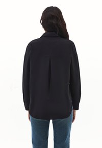 Frau mit langen dunklen Haaren, die ein lockeres schwarzes Langarmhemd und blaue Jeans trägt, steht mit dem Rücken zu einem weißen Hintergrund.