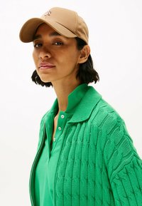 Cardigan verde lavorato a maglia con motivo a trecce, indossato sopra una polo verde con bottoni. Un cappello beige completa l'outfit.