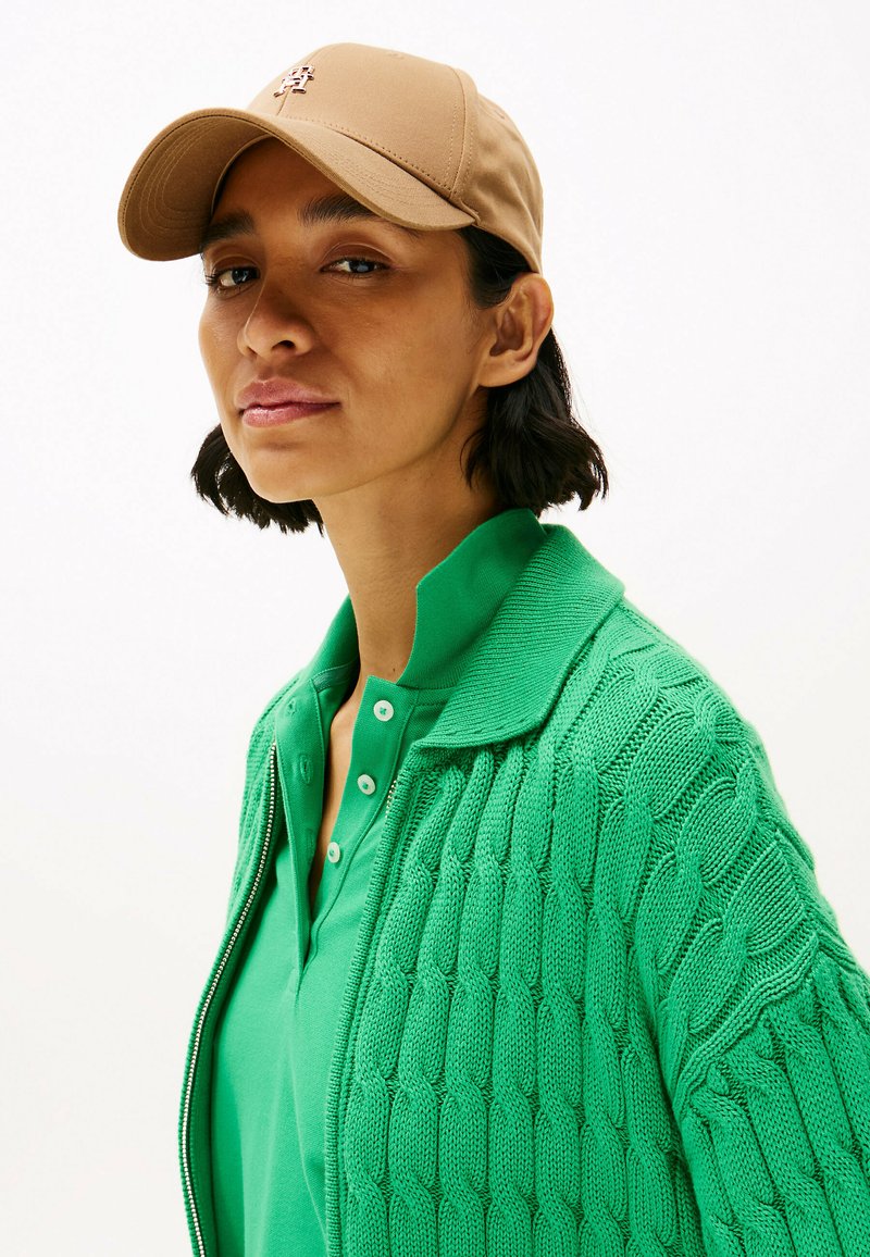 Cardigan verde lavorato a maglia con motivo a trecce, indossato sopra una polo verde con bottoni. Un cappello beige completa l'outfit.