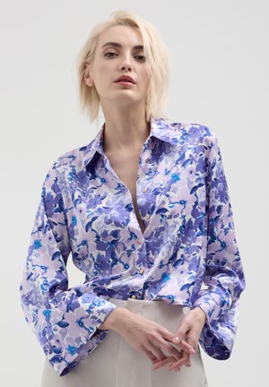 Femme avec des cheveux blonds courts portant un chemisier en satin fleuri violet et un pantalon de couleur claire sur un fond uni.