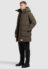 Vycpaný parka kabát olivově zelené barvy s kapucí, zipovou náprsní kapsou, bočními kapsami a zdrsněnými manžetami, nošený přes tmavé džíny a černé boty.