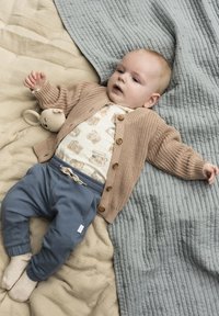 Beige gestrickter Cardigan mit Holzknöpfen, cremefarbenes Shirt mit Kameradrucken, blaue Hose und ein Plüschspielzeug auf einer strukturierten, hellen Decke.