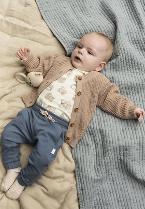 Beige gestrickter Cardigan mit Holzknöpfen, cremefarbenes Shirt mit Kameradrucken, blaue Hose und ein Plüschspielzeug auf einer strukturierten, hellen Decke.