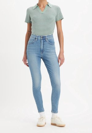 Camisa polo de manga corta a rayas en verde y blanco con cuello en V, combinada con vaqueros ajustados de cintura alta en azul claro y zapatillas blancas.