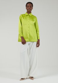 Camisa verde satén de gran tamaño con cuello puntiagudo, dos bolsillos frontales y cierre de botones, combinada con pantalones anchos blancos y tacones transparentes.