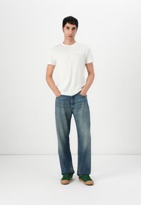Bijela pamučna majica s malim logotipom, plave traperice ravnog kroja i zelene tenisice s smešnim gumastim đonovima. Minimalistički ležeran outfit.