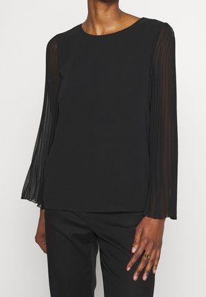 Schwarze Langarmbluse mit transparenten, plissierten Ärmeln, getragen zu schwarzen Hosen, mit rundem Ausschnitt und einem goldenen Ring an der linken Hand.