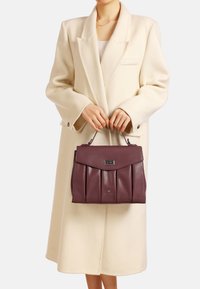 Borsa a mano in pelle bordeaux con design plissettato, manico piatto e accessori in argento, tenuta da una persona in un cappotto bianco.