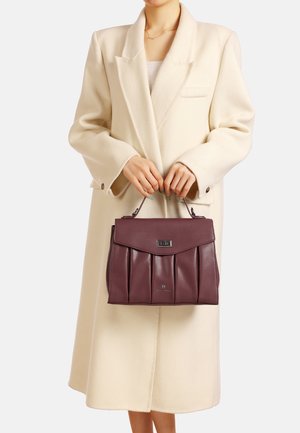 Borsa a mano in pelle bordeaux con design plissettato, manico piatto e accessori in argento, tenuta da una persona in un cappotto bianco.