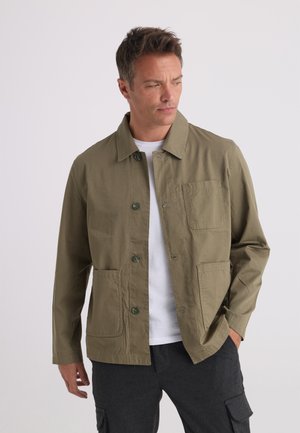 Uomo con giacca verde oliva abbottonata sopra camicia bianca e pantaloni cargo scuri, guarda di lato con una mano in tasca.