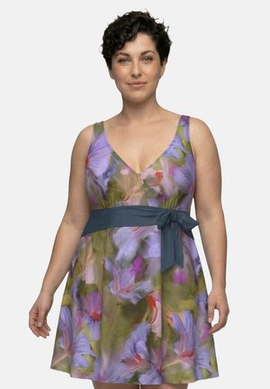 Donna con capelli corti e ricci che indossa un abito floreale senza maniche con tonalità viola e verdi e una cintura blu scuro in vita, in piedi davanti a uno sfondo semplice.