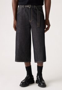 Culottes de mezclilla oscura con pierna ancha, que incluyen un cinturón, costuras amarillas y un ajuste casual. Combinados con calcetines negros y zapatos negros lisos.