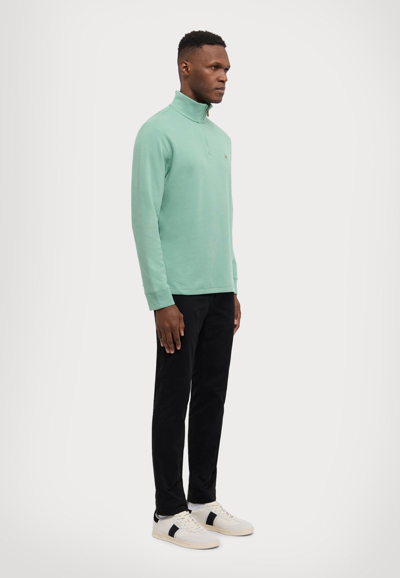 Pullover vert clair à zip quart, avec des manches longues, des poignets côtelés et un petit logo. Associé à un pantalon noir et des baskets blanches.