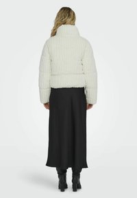 Giacca puffer bianca a coste con colletto alto, abbinata a una lunga gonna nera e stivali neri fino al ginocchio. Il tessuto presenta un aspetto morbido e testurizzato.