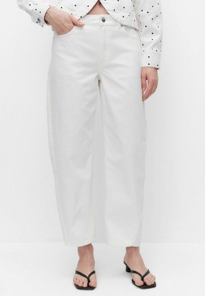 Person iført hvide bukser med vide ben, sorte sandaler med hæl og en hvid skjorte med sorte polkaprikker med lange ærmer, den ene hånd i lommen.