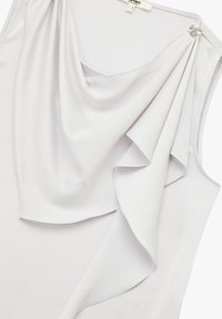 Top sans manches gris clair avec un tissu drapé, une texture douce et un accent décoratif en métal argenté sur une épaule.