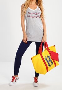 Canottiera grigia senza maniche con lettere rosse, bianche e blu, abbinata a leggings blu scuro e una grande borsa a tracolla gialla con accenti rossi.