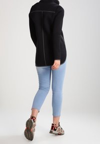 Giacca in pile nera con colletto alto, dettagli in cucitura grigia, abbinata a jeans slim azzurri e scarpe da ginnastica beige con accenti rossi.