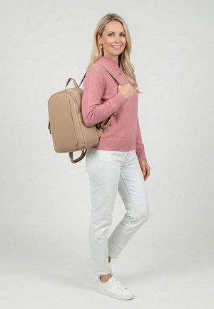 Frau in rosa Pullover und weißer Hose mit einem beigen Rucksack, die lächelt und vor einem schlichten hellgrauen Hintergrund steht.