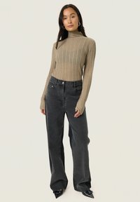 Kvinde iført en beige ribbet turtleneck sweater, vide sorte jeans og sorte spidse hæle, stående mod en ensfarvet lys baggrund.