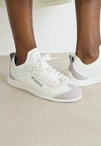 Persona che indossa sneakers bianche e grigie PINKO con colli per caviglia in maglia, in piedi su un pavimento chiaro e liscio.