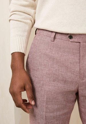 Main droite d'une personne portant un pull en maille beige et un pantalon texturé mauve avec une ceinture boutonnée visible.